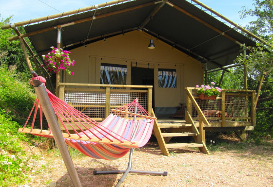 Buitenaanzicht van safaritent Wood Lodge met een kleurrijke hangmat en bloemen op de houten veranda.