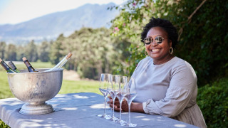Mujer sonriente con gafas de sol junto a una mesa con vino y copas en un glamping de parque vacacional.