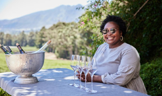 Kvinde smiler med solbriller ved et bord med vin og glas i et naturskønt feriepark-glampingområde.