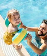 Lille pige med badevinger og mand i swimmingpool ved feriepark med glamping, smiler og hygger sig.
