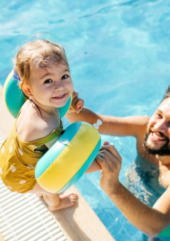 Niña pequeña con flotadores y hombre disfrutando de la piscina en un parque vacacional con glamping.