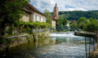 Río pintoresco con casa de piedra, puente y torre de iglesia en valle verde, ideal para glamping.