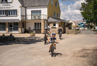 Familie cykler uden for Camping Kost Ar Moor feriepark i Bretagne, Frankrig, på en solrig dag.