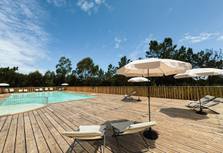 Area piscina soleggiata con lettini e ombrelloni su terrazza in legno al Village Huttopia Lagoa de Óbidos.