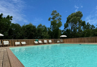 Outdoor pool at Village Huttopia Lagoa de Óbidos, Portugal, with deck chairs and trees under a blue sky.