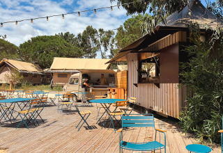 Udendørs caféområde med borde, stole, campinghytter og foodtruck i ferieparken Huttopia Lagoa de Óbidos.