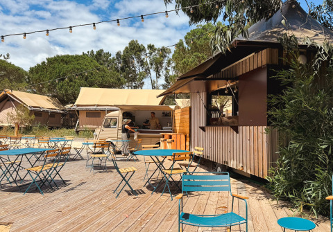 Buitencafé met blauwe tafels, stoelen, foodtruck en tenten bij Huttopia Lagoa de Óbidos vakantiepark, Portugal.