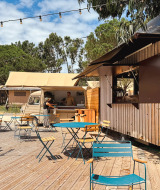 Zona de cafetería al aire libre con mesas, sillas y furgoneta de comida en Huttopia Lagoa de Óbidos, Portugal.