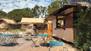 Zona de cafetería al aire libre con mesas, sillas y furgoneta de comida en Huttopia Lagoa de Óbidos, Portugal.
