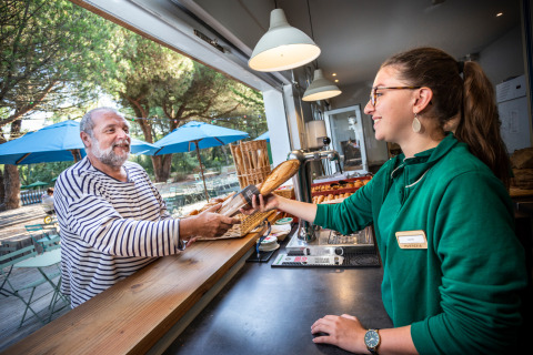 Ein Mann nimmt ein Baguette von einer Mitarbeiterin an einem Café-Schalter im Village Huttopia Lagoa de Óbidos.