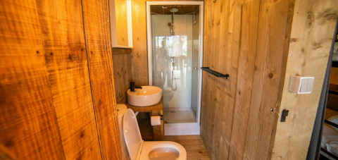 Baño con paredes de madera, inodoro, lavabo y ducha en una tienda Safari Lodge en Camping Kost Ar Moor, Francia.
