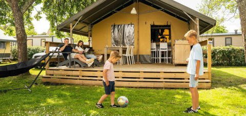 Familia disfrutando en tienda safari lodge, mientras dos niños juegan al balón frente al porche y padres descansan.