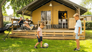Familia disfrutando en tienda safari lodge, mientras dos niños juegan al balón frente al porche y padres descansan.