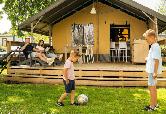 Gezin geniet voor een safari lodge tent, met twee kinderen die voetballen op het gras en ouders op het terras.