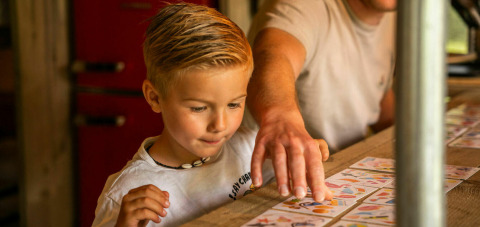 Een jongen en een volwassene spelen samen kaarten aan een tafel in een safaritent op Camping Kost Ar Moor, Frankrijk.