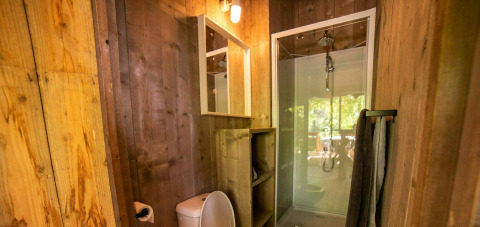 Bagno in tenda safari con pareti in legno, doccia vetro, WC, specchio, scaffali e porta asciugamani.