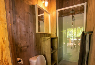 Baño de tienda safari con paredes de madera, ducha de vidrio, inodoro, espejo y toallas colgadas.