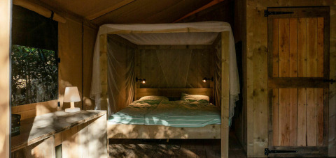 Vista interior de Safari Lodge con cama doble, lámpara de mesa y muebles de madera en Camping Les Tailladis.