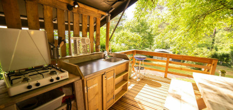 Outdoor kitchen with a gas stove, sink, and wooden deck surrounded by lush greenery in a safari tent.