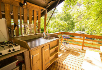 Outdoor kitchen with a gas stove, sink, and wooden deck surrounded by lush greenery in a safari tent.
