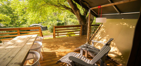 Wooden deck with dining table, chairs and lounge seats outside a safari tent at Camping Les Tailladis, France.