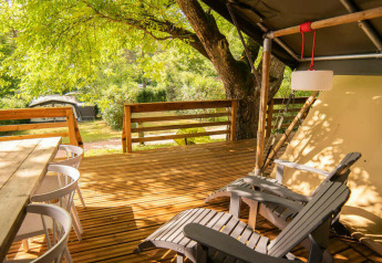 Wooden deck with dining table, chairs and lounge seats outside a safari tent at Camping Les Tailladis, France.