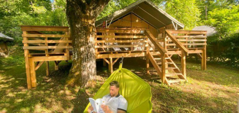 Man reading in a hammock under a tree in front of a wooden cabin at Camping Les Tailladis, Nouvelle-Aquitaine.