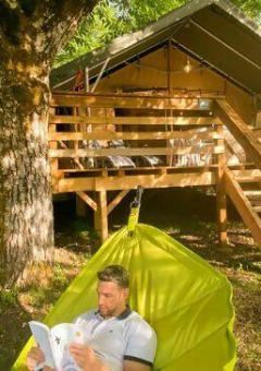 Hombre leyendo en una hamaca bajo un árbol frente a una cabaña en Camping Les Tailladis, Nouvelle-Aquitaine.