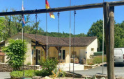 Billede af Camping Les Tailladis med sving, hovedbygning, flag og parkeret bil under en solrig himmel.