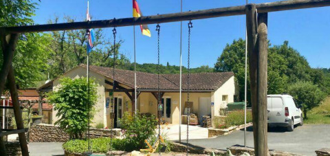 Billede af Camping Les Tailladis med sving, hovedbygning, flag og parkeret bil under en solrig himmel.