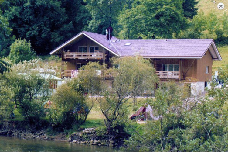 Parc de vacances avec un grand chalet en bois au bord d'une rivière, cadre verdoyant idéal pour glamping.