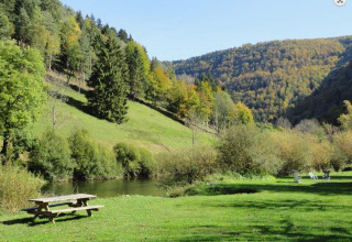Udsigt over frodig dal med picnicbord, græsplæne, en flod og træbeklædte bakker i et feriepark.