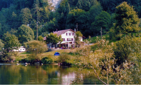 Vakantiepark met glamping langs het water, omgeven door groene heuvels, met tent en huis in beeld.