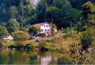 Parque vacacional con glamping junto al lago, rodeado de bosques y colinas, con tienda y casa visibles.