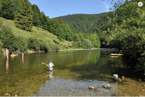 Niños y adultos juegan en un río poco profundo junto a un parque de glamping rodeado de colinas verdes.