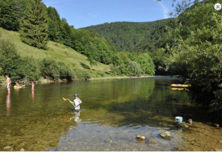 Niños y adultos juegan en un río poco profundo junto a un parque de glamping rodeado de colinas verdes.