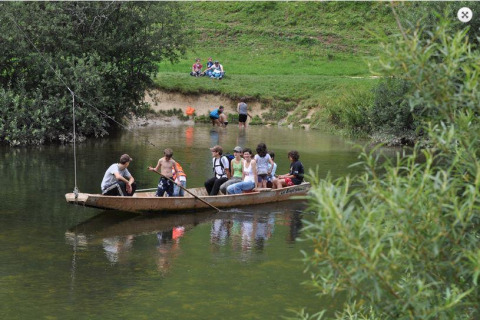 Børn og voksne sejler i en lille båd på en flod i en feriepark med grønne områder og glamping.