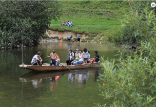 Børn og voksne sejler i en lille båd på en flod i en feriepark med grønne områder og glamping.