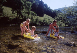 Niños juegan en botes inflables sobre un río poco profundo, rodeados de naturaleza en un parque de glamping.
