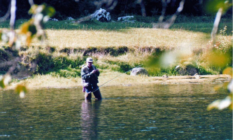 Man vliegvist in een rivier bij een glampingvakantiepark, omgeven door natuur en zonnig grasveld.