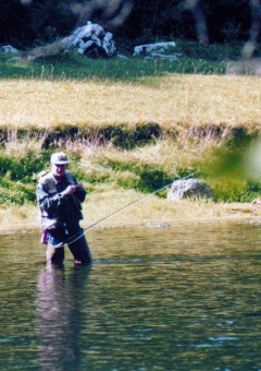 Homme faisant de la pêche à la mouche dans une rivière d’un parc de glamping, entouré de verdure.