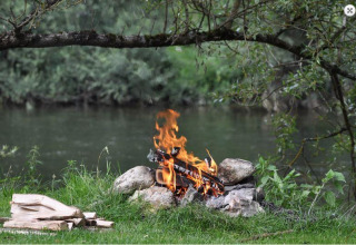 Kampvuur aan het water, omringd door groene natuur, voor gezellige avonden bij het glampen.