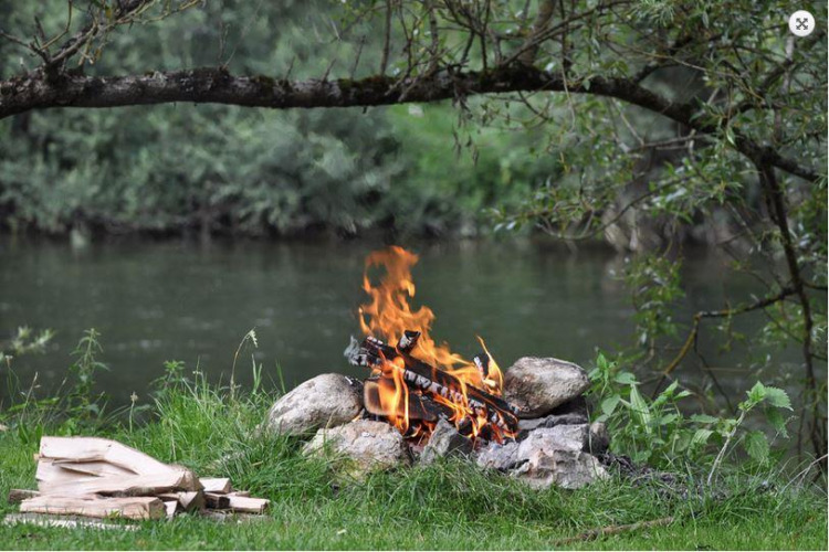Feu de camp au bord de la rivière entouré de verdure, parfait pour une soirée détente en glamping.