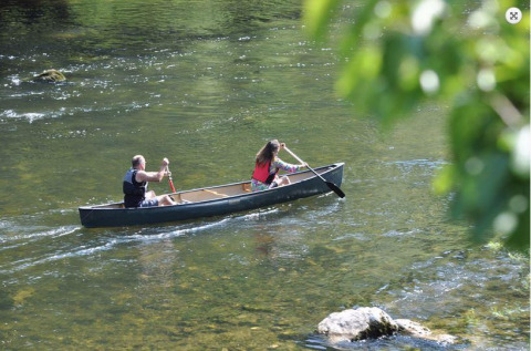 To personer padler i en kano på en rolig flod ved en feriepark med glamping, omgivet af grøn natur.