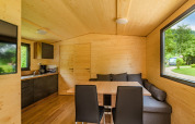 Moderne træhytteinteriør med spisebord, hjørnesofa, stort vindue og køkken i Drau Camping Sachsenburg.
