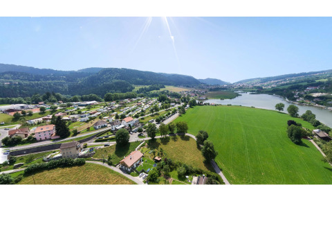Vista aérea del Camping Lac des Brenets en Neuchâtel, Suiza, mostrando campos verdes, río y colinas al fondo.