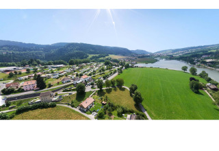 Vista aérea del Camping Lac des Brenets en Neuchâtel, Suiza, mostrando campos verdes, río y colinas al fondo.