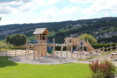 Parque infantil de madera en Camping Lac des Brenets, parque de vacaciones en Neuchâtel, Suiza, con colinas al fondo.