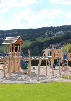 Parque infantil de madera en Camping Lac des Brenets, parque de vacaciones en Neuchâtel, Suiza, con colinas al fondo.