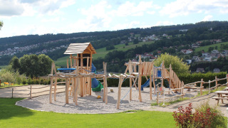 Parque infantil de madera en Camping Lac des Brenets, parque de vacaciones en Neuchâtel, Suiza, con colinas al fondo.